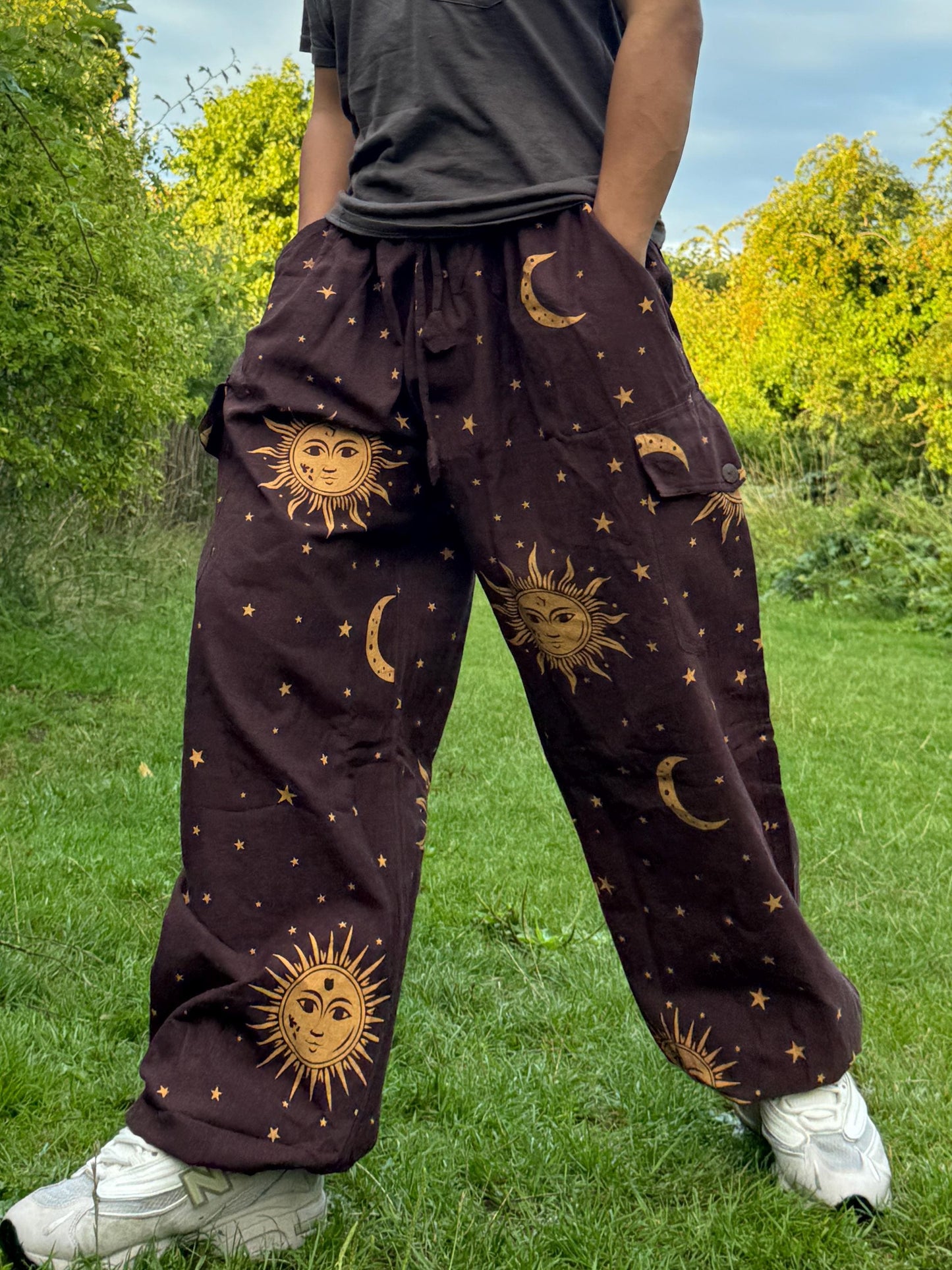 May include: Brown trousers with a celestial design featuring suns, moons, and stars. The trousers have a relaxed fit with pockets and a drawstring waist. The sun and moon graphics are in a golden colour, creating a striking contrast against the brown fabric.