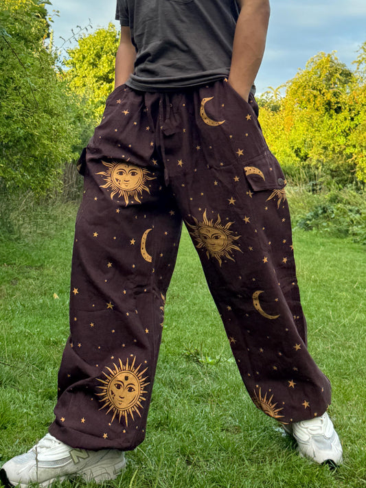 May include: Brown trousers with a celestial design featuring suns, moons, and stars. The trousers have a relaxed fit with pockets and a drawstring waist. The sun and moon graphics are in a golden colour, creating a striking contrast against the brown fabric.