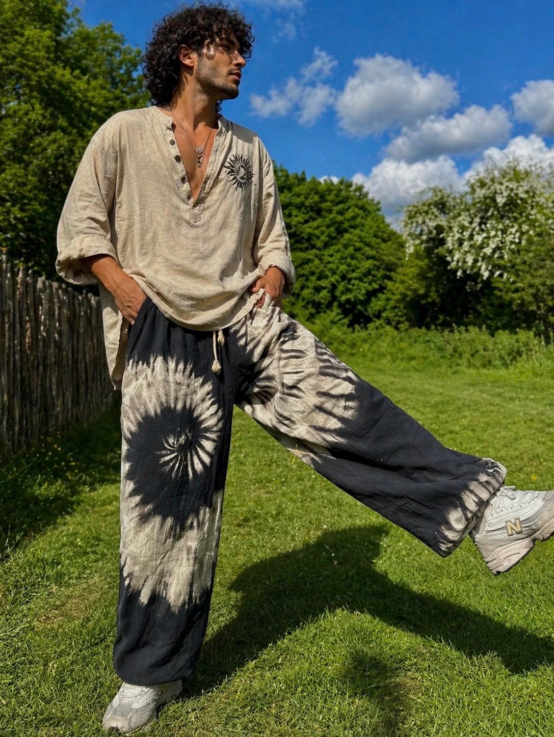 May include: A beige long-sleeved shirt with a sun graphic and black and white tie-dye trousers. The trousers feature a spiral design. The person is wearing grey trainers and standing on green grass.